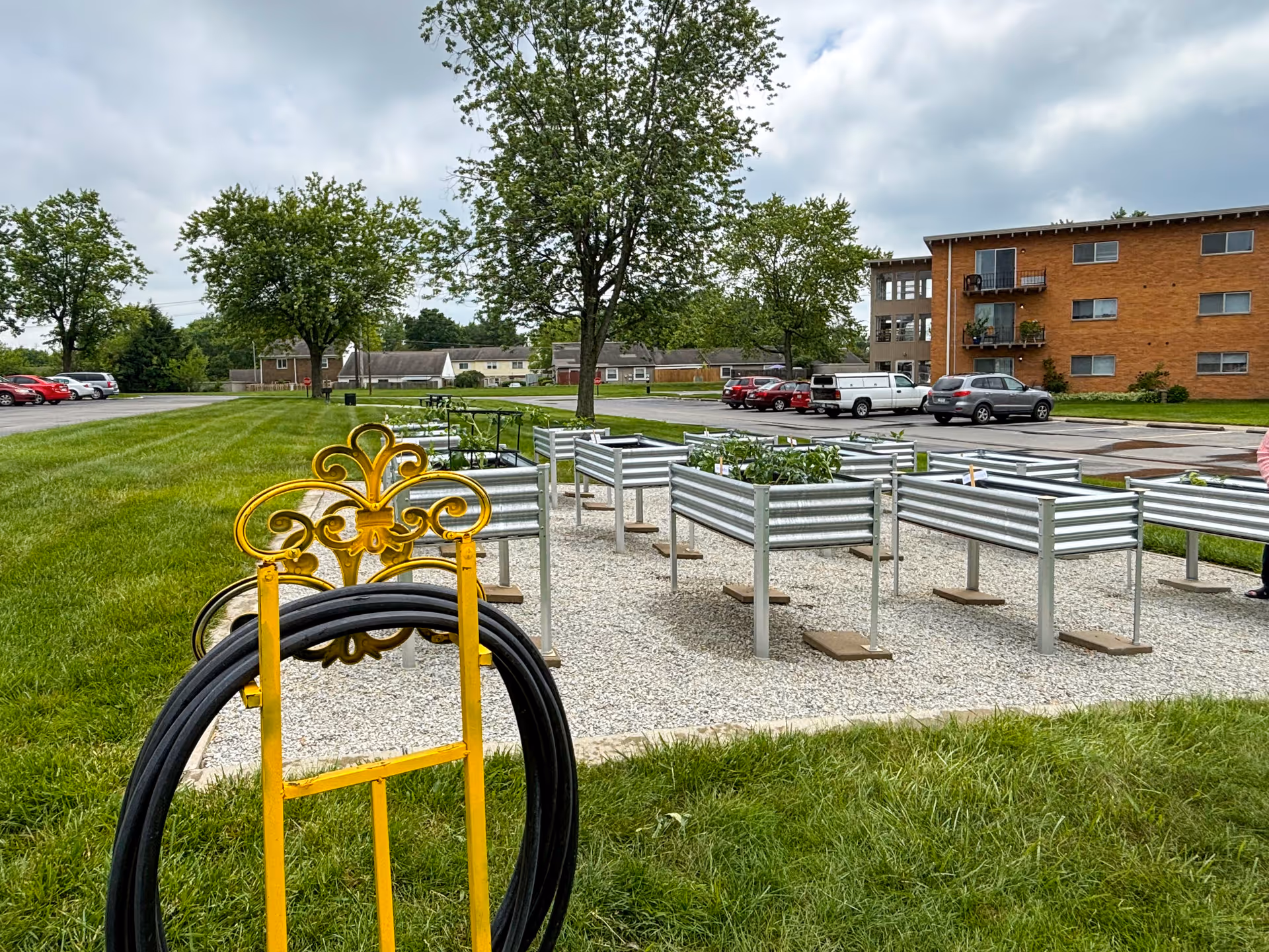 Outdoor garden area with multiple raised garden beds filled with plants, surrounded by gravel and grass. A yellow decorative metal stand with a coiled black hose is in the foreground. Trees, parked cars, and a multi-story brick building are visible in the background under a cloudy sky.