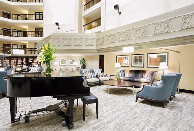 A spacious and elegant living room area with a black grand piano in the foreground, surrounded by comfortable seating including armchairs and sofas. The room features a high ceiling with multiple balconies overlooking the space, decorative wall art, lamps, and a bookshelf in the background.