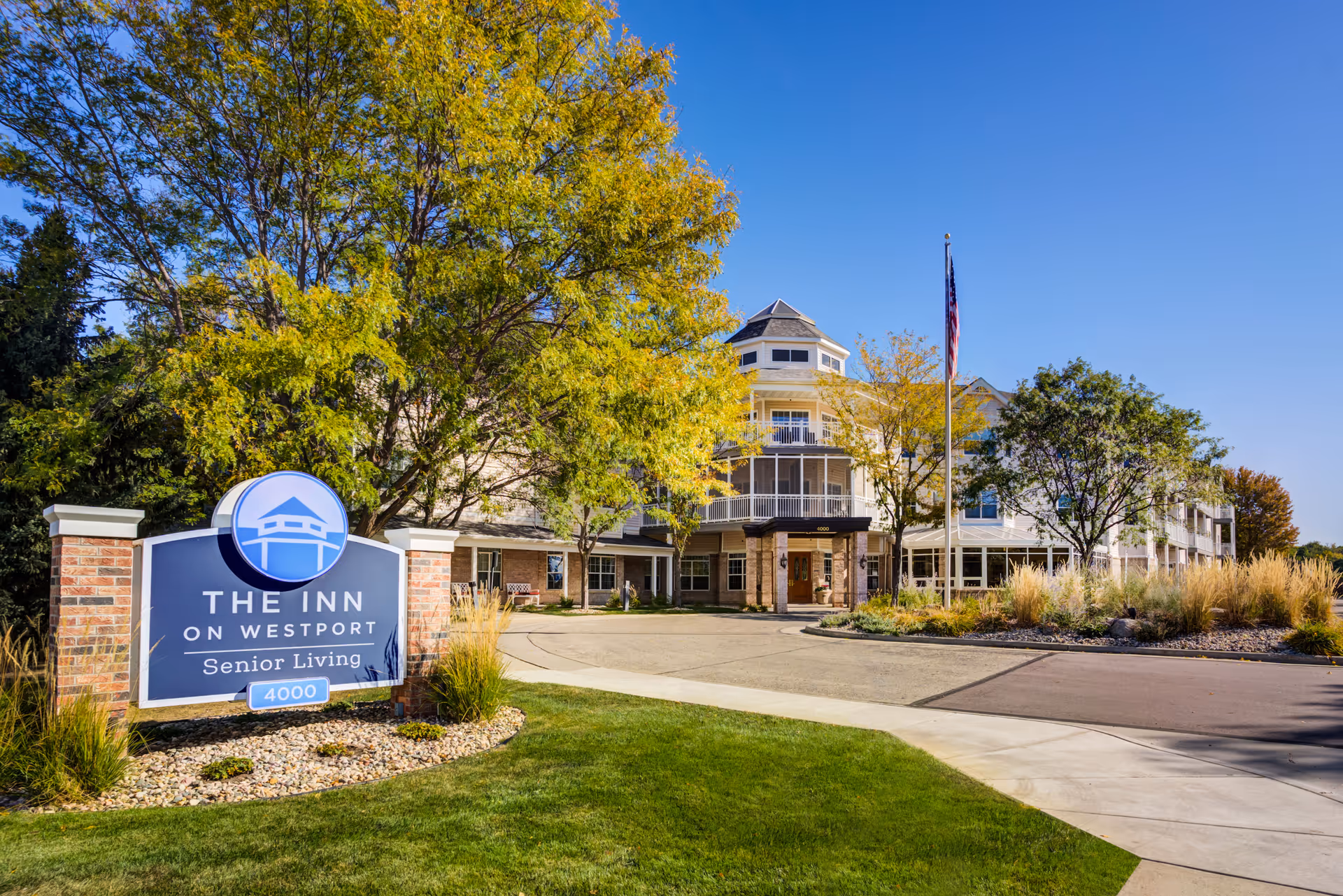 Exterior view of The Inn on Westport senior living facility with a clear blue sky, trees with green and yellow leaves, a driveway, and an American flag on a flagpole.