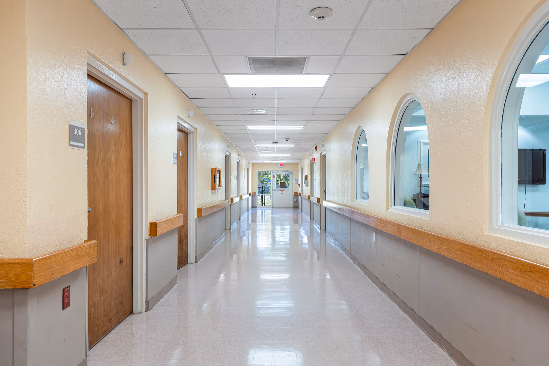 A clean, well-lit hallway in a senior living facility with beige walls, wooden handrails, and several closed wooden doors on the left side. On the right side, there are arched windows looking into a room with furniture. The floor is shiny and reflective, and the ceiling has recessed lighting panels.