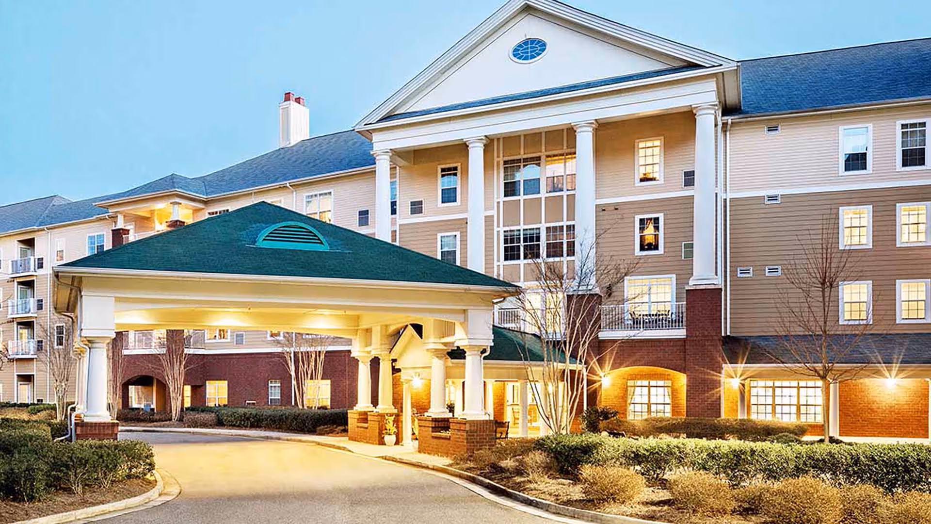 Exterior view of Atria North Point senior living facility at dusk, showing a large building with multiple windows, a covered entrance with white columns, and well-maintained landscaping.