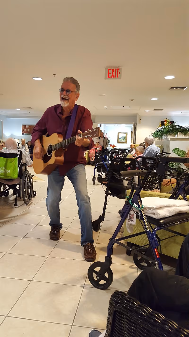 An elderly man with gray hair and beard is playing an acoustic guitar and singing in a common area of an assisted living facility. Several elderly residents are seated around the room, some in wheelchairs and others in chairs, watching and enjoying the performance. The room has tiled floors, white walls, and ceiling lights, with plants and framed pictures decorating the space.