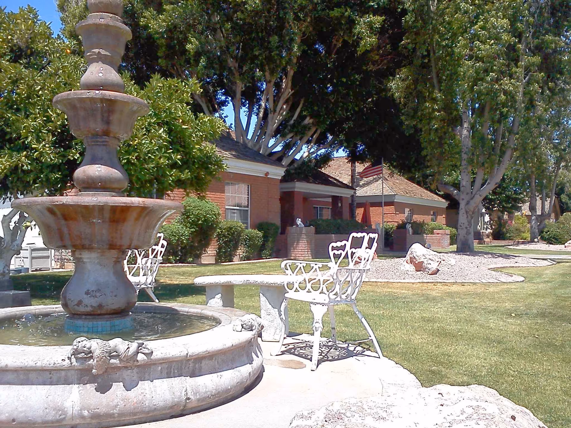 Outdoor garden area with a large stone fountain in the foreground, white metal chairs and a stone table nearby, green grass, trees, and a brick building in the background with an American flag on a flagpole.