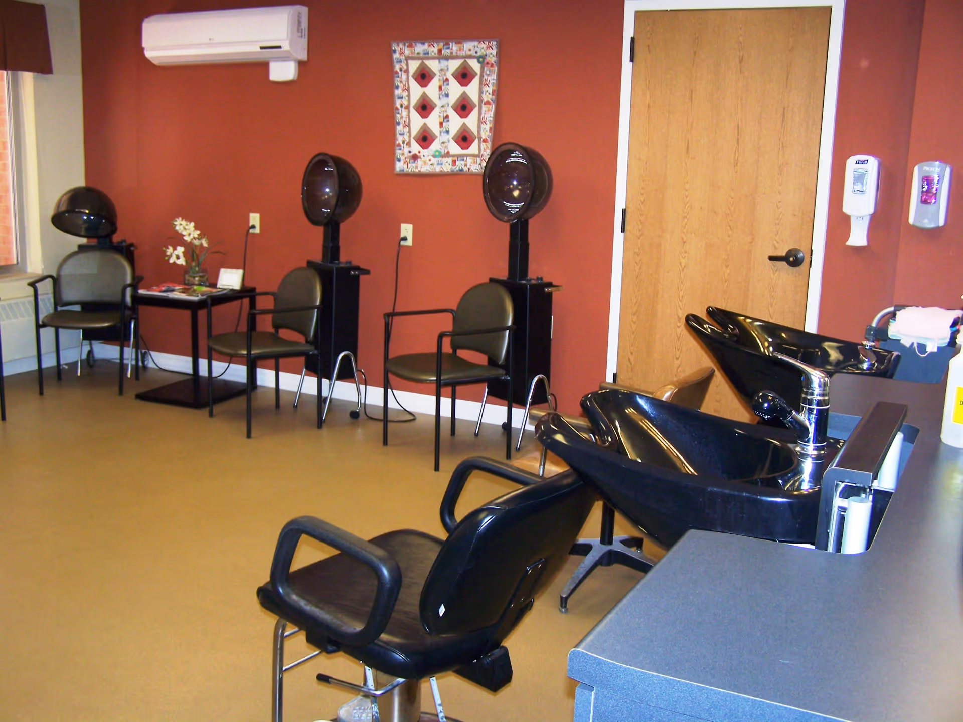 Interior of a hair salon area with three black hair drying chairs against a red wall, two black hair washing sinks with chairs in front, a small table with flowers and magazines, and a wooden door. The room has a beige floor and an air conditioning unit mounted on the wall.