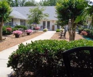 Outdoor courtyard area at a senior living facility with a paved walkway, green bushes, trees, benches, and a building with multiple doors and windows in the background.