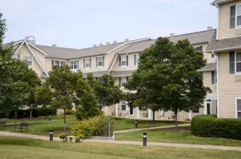 A beige multi-story senior living building with green lawn and trees.