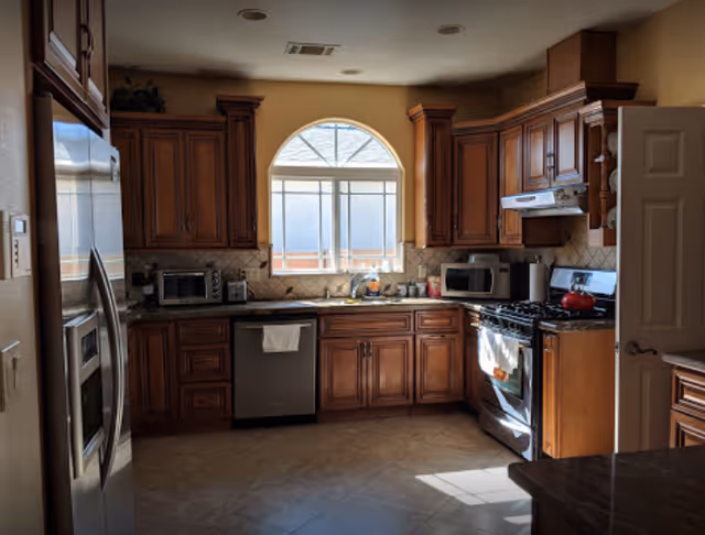 Sunlit kitchen with wooden cabinets, stainless steel refrigerator and dishwasher, gas stove, microwave, and an arched window above the sink.