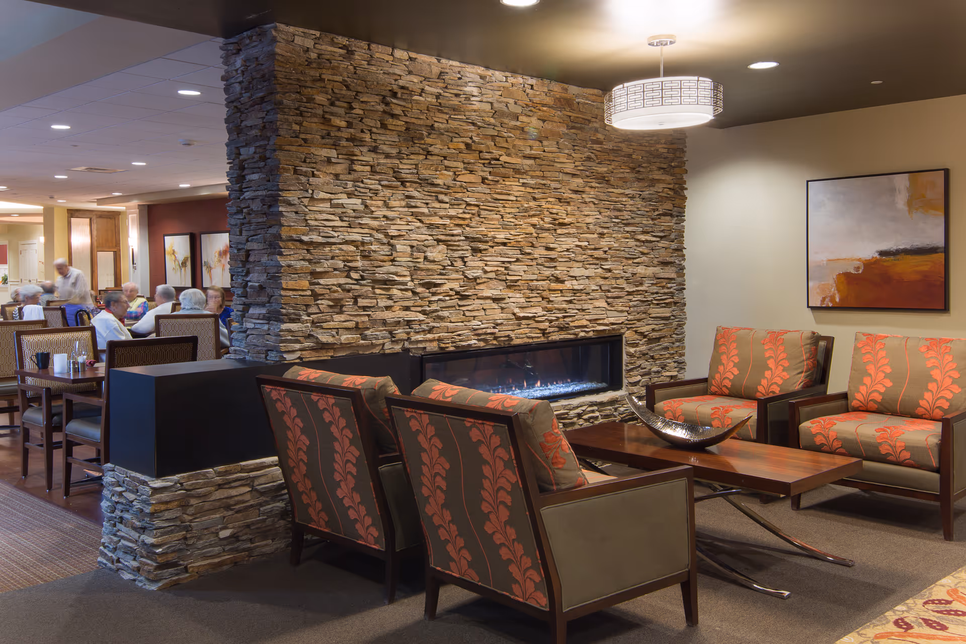 A cozy senior living facility common area with a stone fireplace in the center. The seating area includes four armchairs with red and brown patterned upholstery arranged around a wooden coffee table with a decorative bowl. In the background, there is a dining area with several elderly people seated at tables. The walls are decorated with modern artwork and the space is warmly lit.