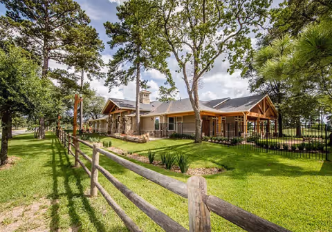 Exterior view of Village Green Memory Care Community Tomball showing a single-story building with a wooden fence in the foreground, surrounded by green grass and tall trees under a partly cloudy sky.