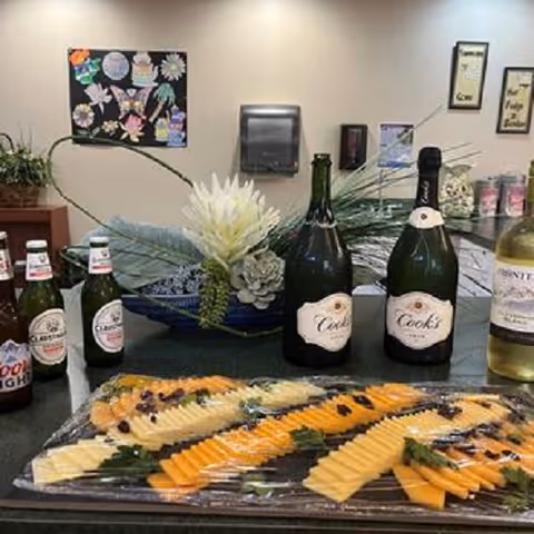 A countertop with a platter of sliced cheese arranged in rows, covered with plastic wrap. Behind the platter are several bottles of beer and wine. In the background, there is a decorative plant arrangement, a paper towel dispenser, and wall decorations including framed quotes and a colorful bulletin board.