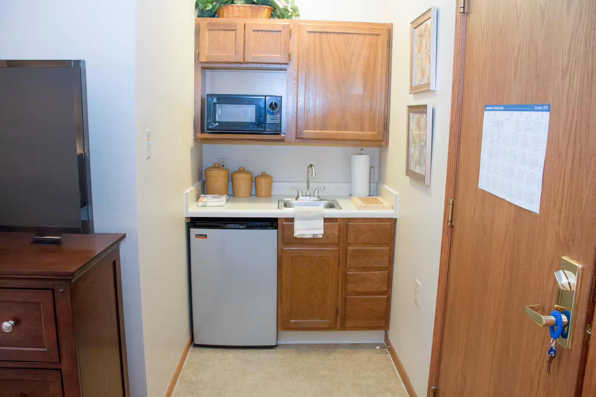Small kitchenette area with wooden cabinets, a microwave, a mini refrigerator, a sink with a towel hanging on it, three ceramic canisters, a paper towel holder, and a cutting board. To the right is a wooden door with a calendar attached, and to the left is a dark wooden dresser with a television on top.