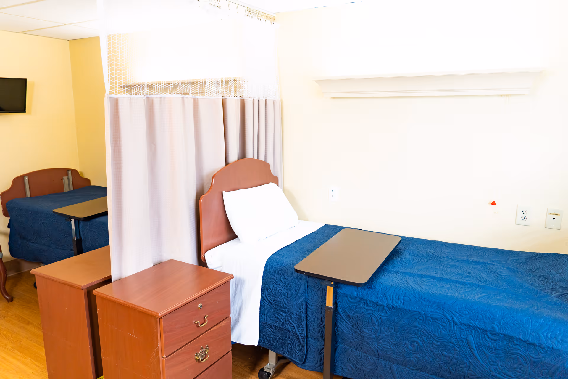 A room with two single beds separated by a privacy curtain. Each bed has a wooden headboard, white pillow, and blue quilted blanket. There is a small wooden nightstand with drawers between the beds, and a small overbed table on wheels is positioned over one of the beds. A wall-mounted TV is visible in the background.