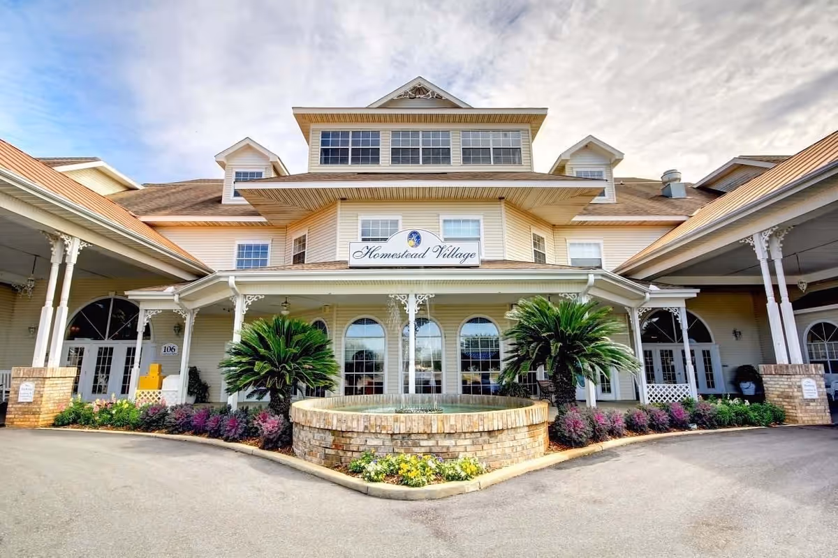 Front exterior view of Homestead Village Of Fairhope, a large building with a covered entrance, decorative columns, a circular brick planter with greenery, and a sign displaying the facility name.