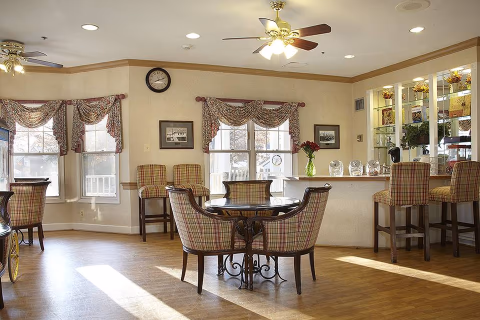 A bright and cozy common area with wooden flooring, plaid upholstered chairs around a round glass table, and a counter with bar stools. The room has large windows with floral valances, a ceiling fan with lights, a clock on the wall, and decorative items on shelves behind the counter.