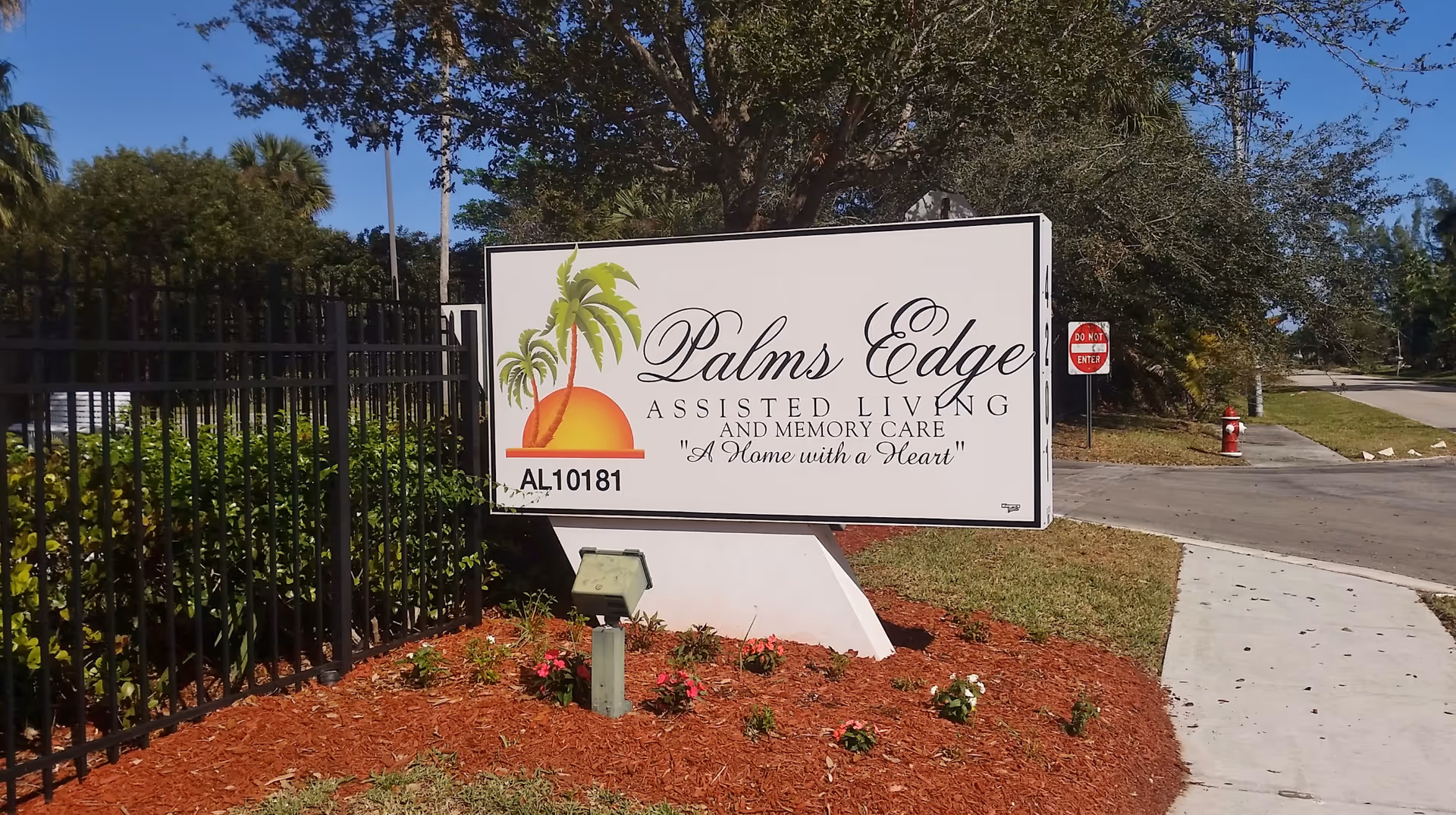 Outdoor view of a sign for Palms Edge Assisted Living and Memory Care, featuring a logo with palm trees and a sunset. The sign is placed in a landscaped area with mulch and flowers, next to a black metal fence and a sidewalk. A 'Do Not Enter' sign and a fire hydrant are visible in the background along the street.