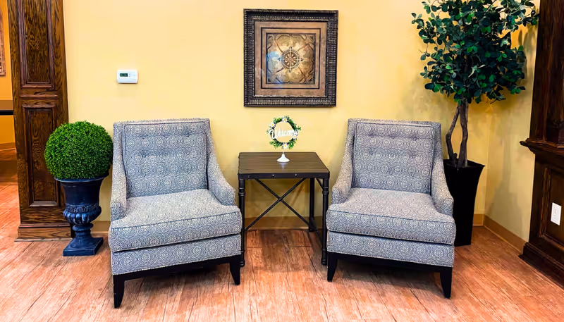 Two patterned armchairs with dark wooden legs are positioned on either side of a small dark wooden table with a decorative 'Welcome' sign on it. Behind the chairs is a yellow wall with a framed artwork hanging in the center. To the left of the left chair is a round green topiary plant in a black decorative pot, and to the right of the right chair is a taller leafy green plant in a black pot. The floor is wooden with a warm tone.