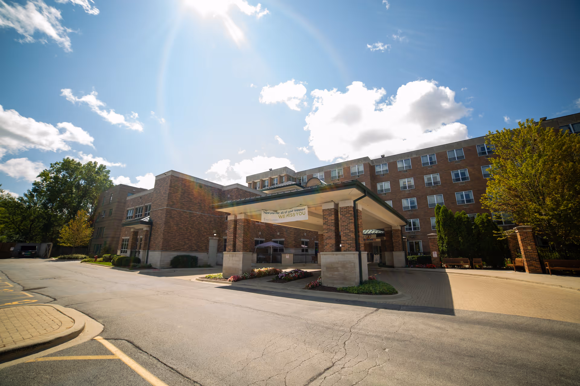 Exterior view of a multi-story brick building with a covered entrance and a driveway in front. The sky is blue with some clouds, and there are trees and bushes around the building. A banner hangs from the covered entrance.