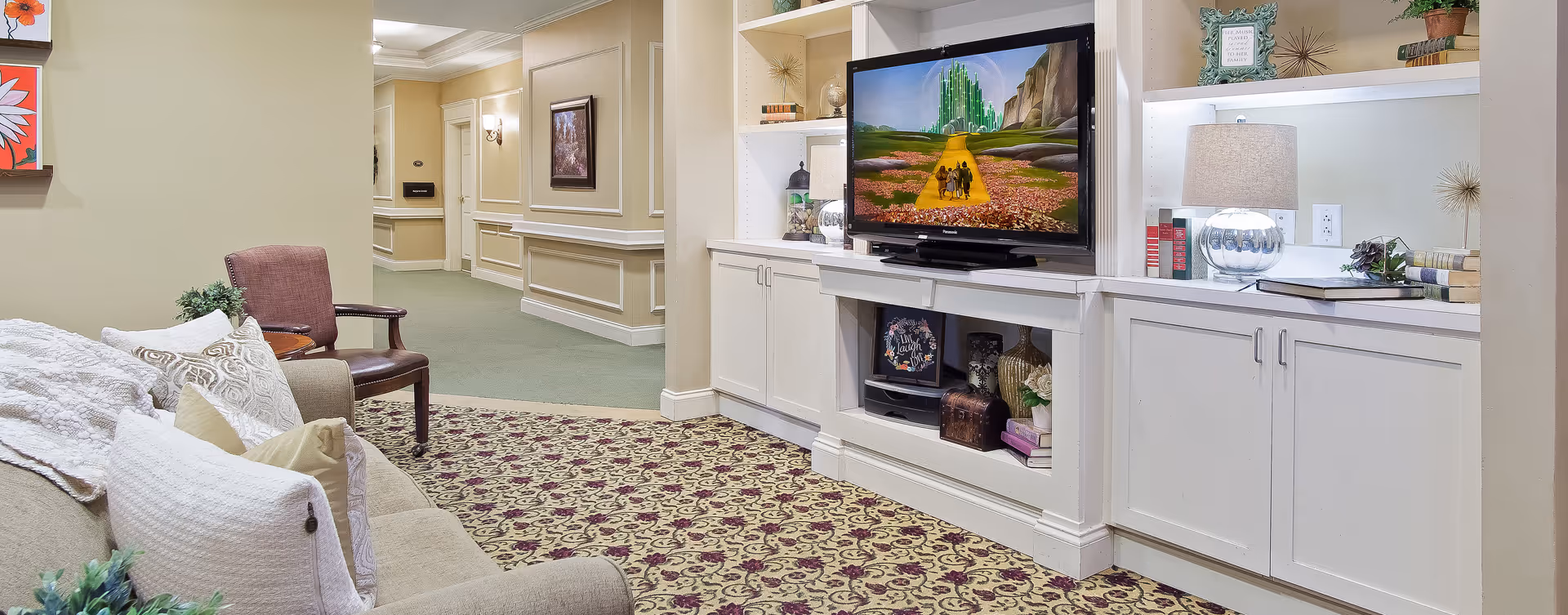 Common sitting area with a sofa and armchair facing a built-in TV and shelving in a senior living facility corridor.