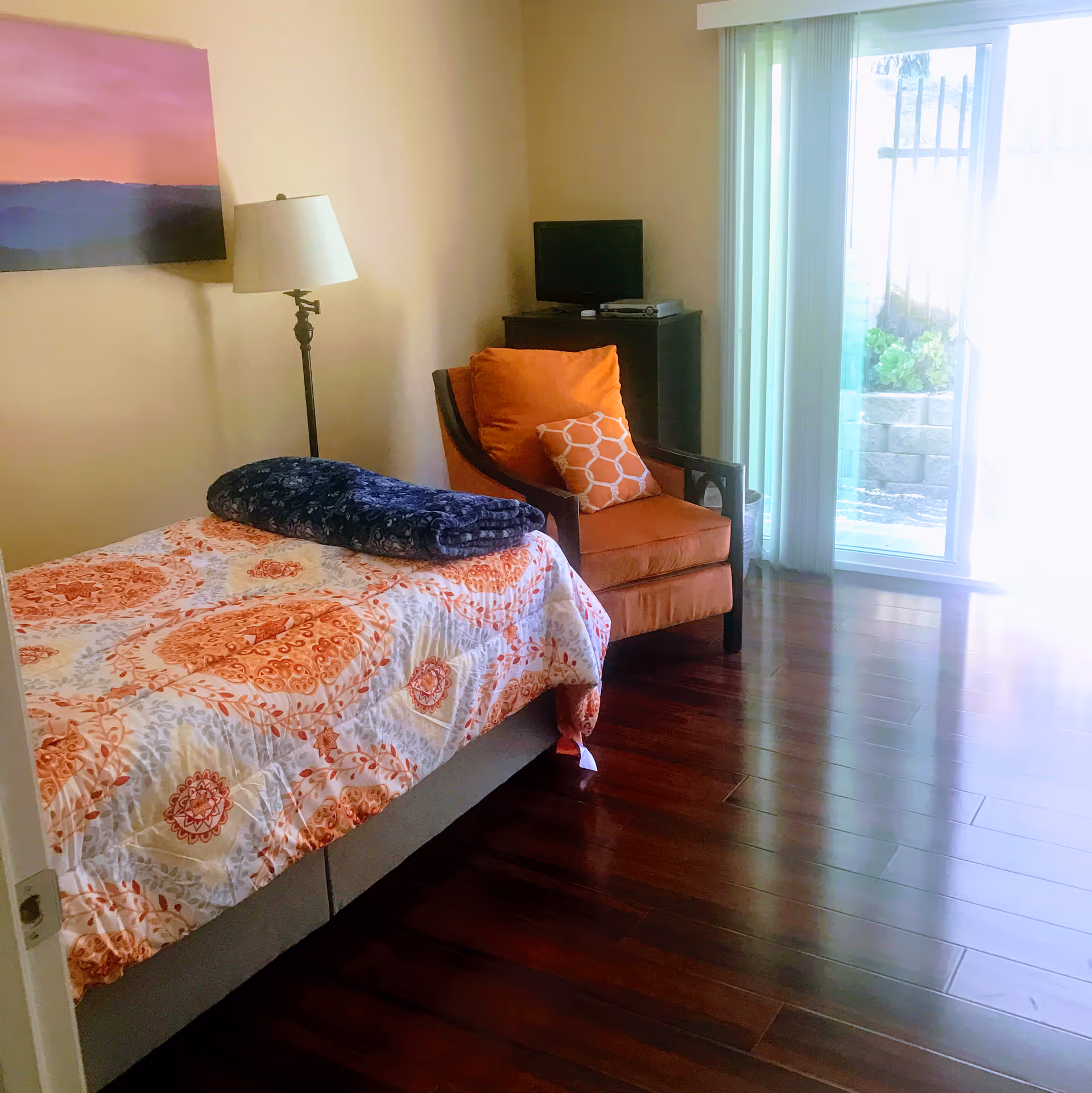 A bedroom with a single bed covered in a patterned orange and white quilt with a folded dark blue blanket on top. Next to the bed is an orange armchair with two decorative pillows. A floor lamp stands beside the bed, and a small TV sits on a dark wooden cabinet in the corner. The room has wooden flooring and a sliding glass door with vertical blinds leading outside.