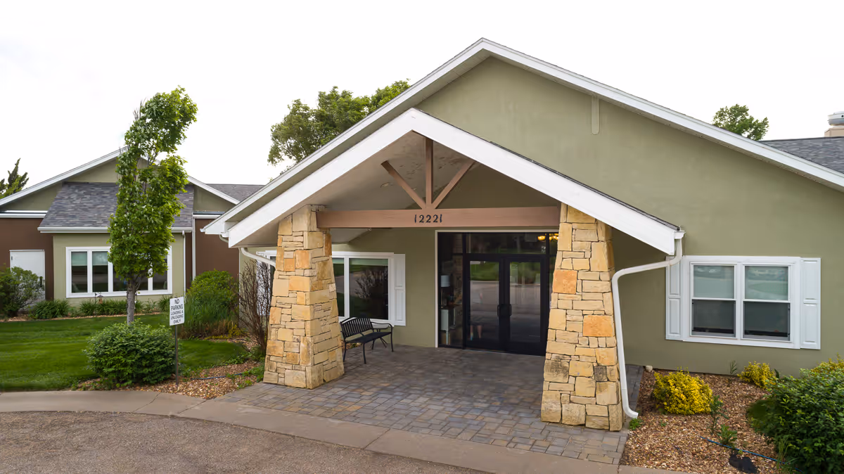 Covered front entrance of a single-story assisted living building with stone pillars, double glass doors, and landscaped grounds.