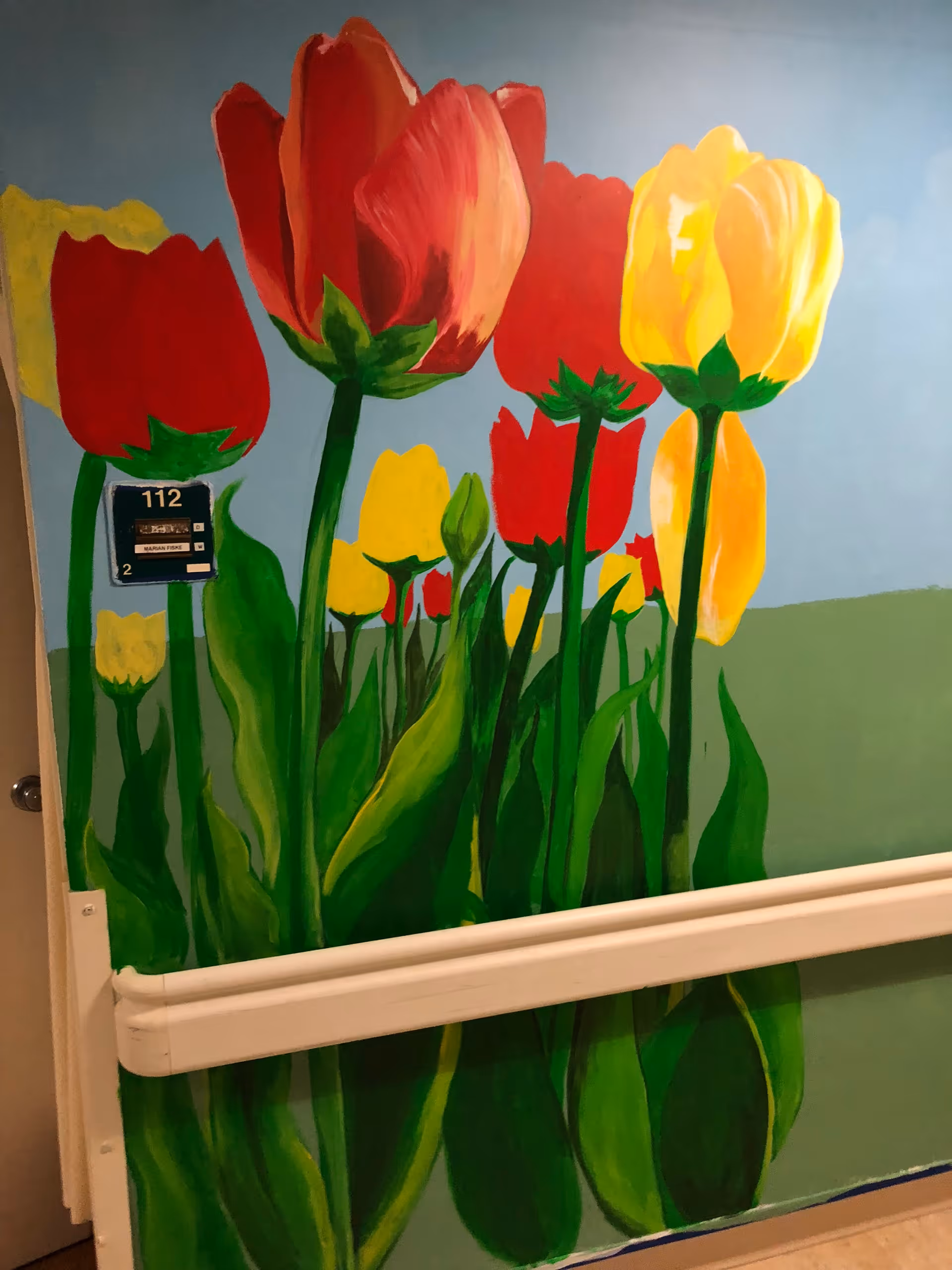A colorful mural painted on a wall depicting large red and yellow tulips with green stems and leaves against a blue sky and green grass background. A room number plaque labeled '112' is visible on the wall.