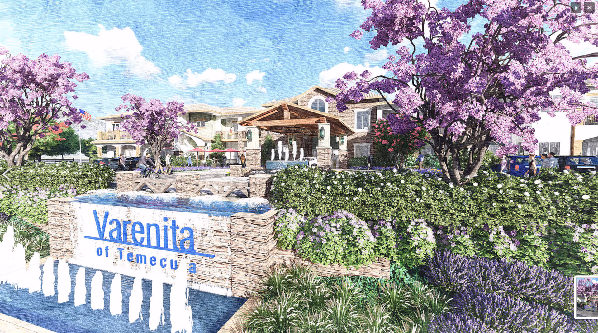 Exterior view of Varenita of Temecula senior living facility with a stone sign and water fountain in the foreground, surrounded by lush greenery and purple flowering trees. The building features a covered entrance with stone pillars and balconies, with people walking and cars parked nearby under a partly cloudy sky.