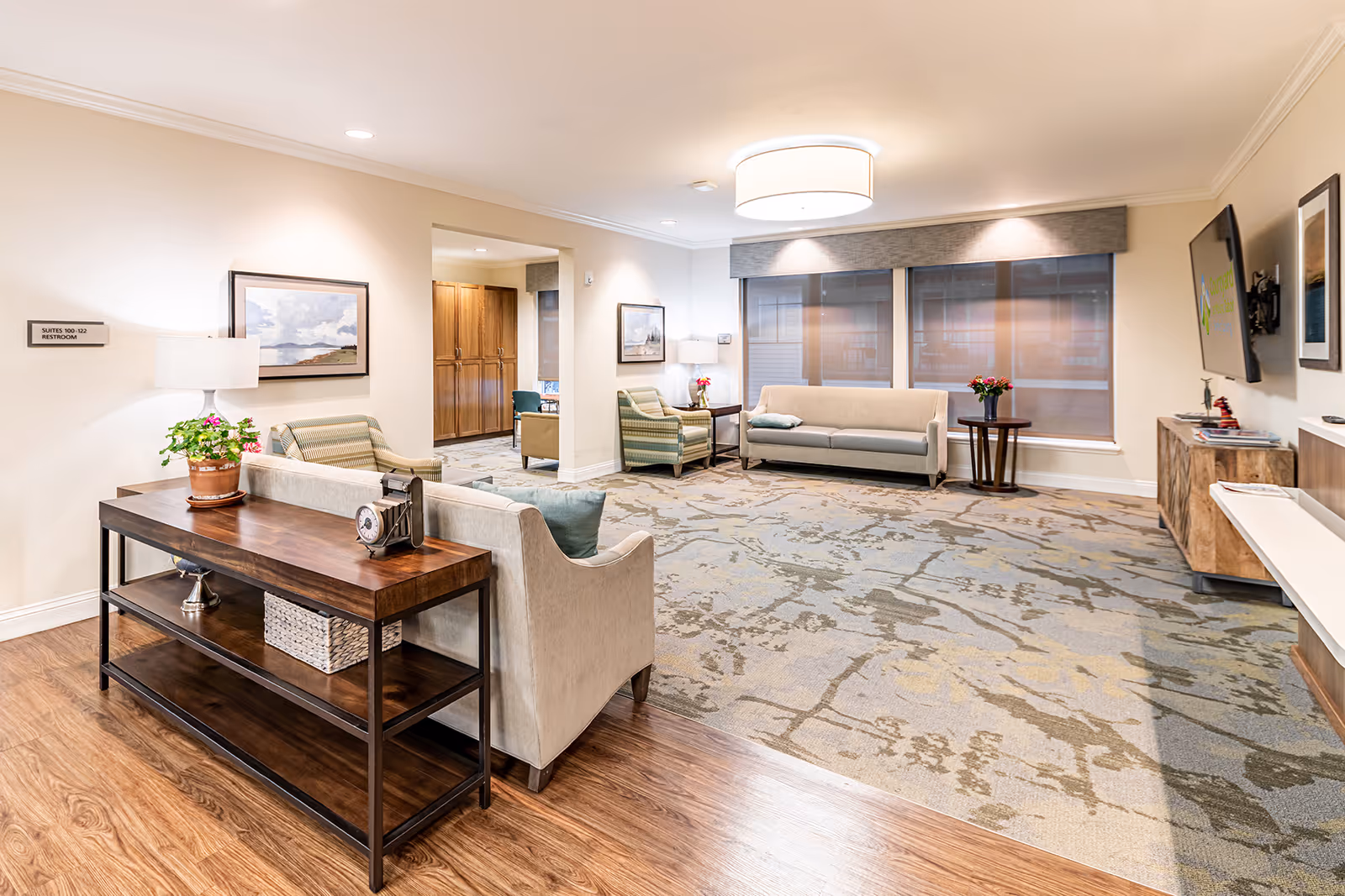A bright and spacious living room area in a senior living facility with beige sofas, striped armchairs, a wooden console table with a potted plant and decorative clock, a large patterned carpet, and a wall-mounted TV. Large windows with shades allow natural light to fill the room, and framed artwork decorates the walls.