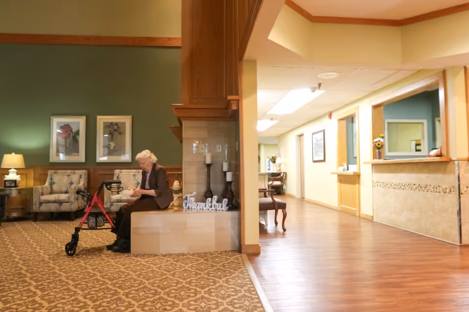An elderly woman sitting on a bench in a senior living facility lobby area with a red walker in front of her. The room has patterned carpet, green walls with framed floral artwork, and comfortable armchairs. To the right is a hallway with wooden flooring leading to a reception desk with a small flower vase and a window opening to the office area. There are decorative candles and a 'Thankful' sign on the bench beside the woman.