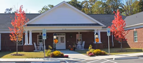 Front exterior view of a single-story brick building with a covered entrance supported by white columns. There are two red-leaved trees on either side of the entrance, rocking chairs on the porch, and handicap parking signs visible in front.