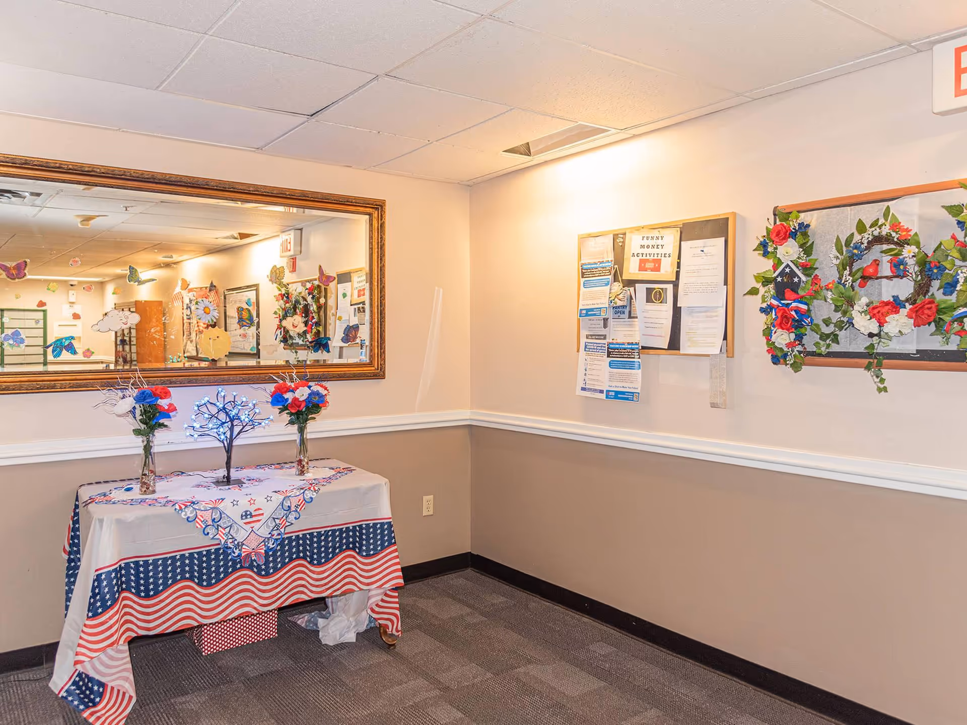Interior corner of a senior living facility hallway with a table covered in a patriotic-themed tablecloth, two vases with red, white, and blue flowers, and a small decorative lighted tree. A large mirror on the left wall reflects part of the hallway decorated with butterfly and flower decals. On the right wall, there is a bulletin board with various notices and a decorative wreath with red, white, and blue flowers and greenery.