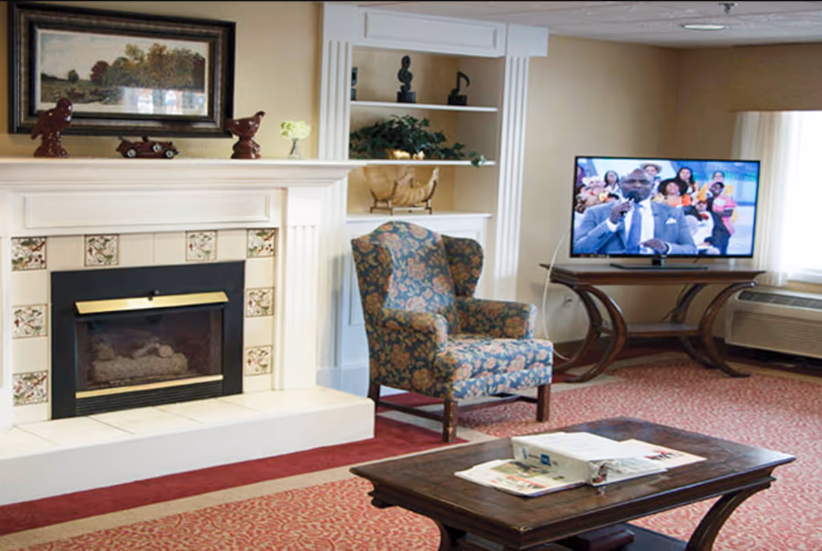 A cozy living room with a floral upholstered armchair, a wooden coffee table with newspapers and a binder, a fireplace with decorative tiles and a mantel holding small sculptures and a framed picture. A flat-screen TV on a wooden stand shows a man speaking to an audience. The room has beige walls, a red patterned carpet, and a window with white curtains.