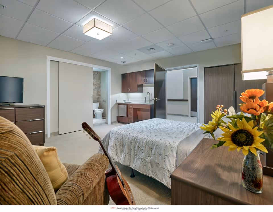 A senior living facility bedroom with a bed covered in a white patterned bedspread, a brown armchair with a yellow pillow, a guitar resting against the armchair, a wooden dresser with a flat-screen TV, and a wooden nightstand with a vase of sunflowers and a lamp. The room has a kitchenette area with a sink and cabinets, and an open door leading to a bathroom with a visible toilet.