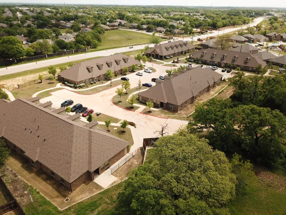 Aerial view of Mayberry Gardens Assisted Living and Memory Care Homes showing multiple single-story buildings with brown roofs arranged around a parking area with several cars. The facility is surrounded by green trees and residential neighborhoods in the background.