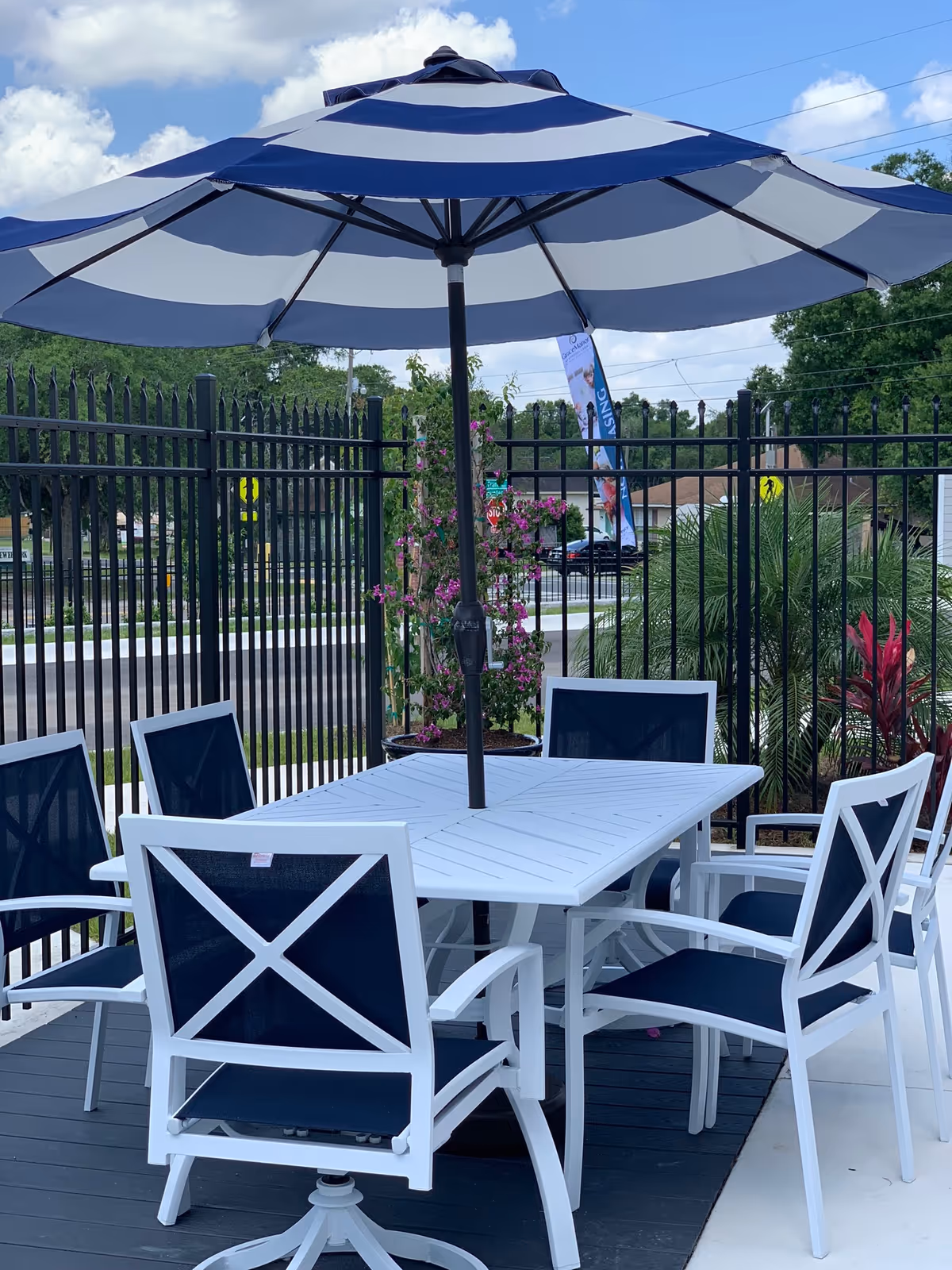 Outdoor patio area with a white table and six white chairs with dark blue seats and backs, shaded by a large blue and white striped umbrella. The area is enclosed by a black metal fence, with greenery and a clear blue sky in the background.