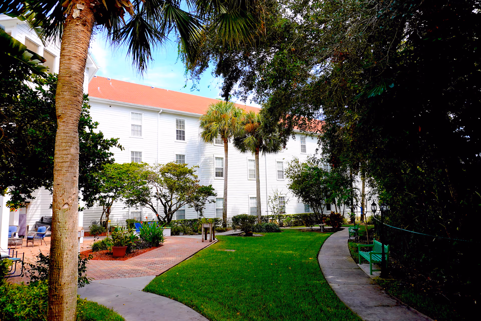Outdoor garden area at Sodalis of Largo featuring a curved concrete walkway, green grass, palm trees, and other foliage. There are green benches along the path and a white multi-story building with a red roof in the background.