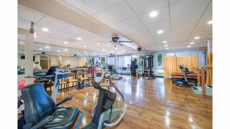 A spacious fitness room with various exercise equipment including stationary bikes, treadmills, and weight machines. The room has wooden flooring, large windows with curtains allowing natural light, ceiling fans, and recessed lighting. Several chairs are arranged along the walls.