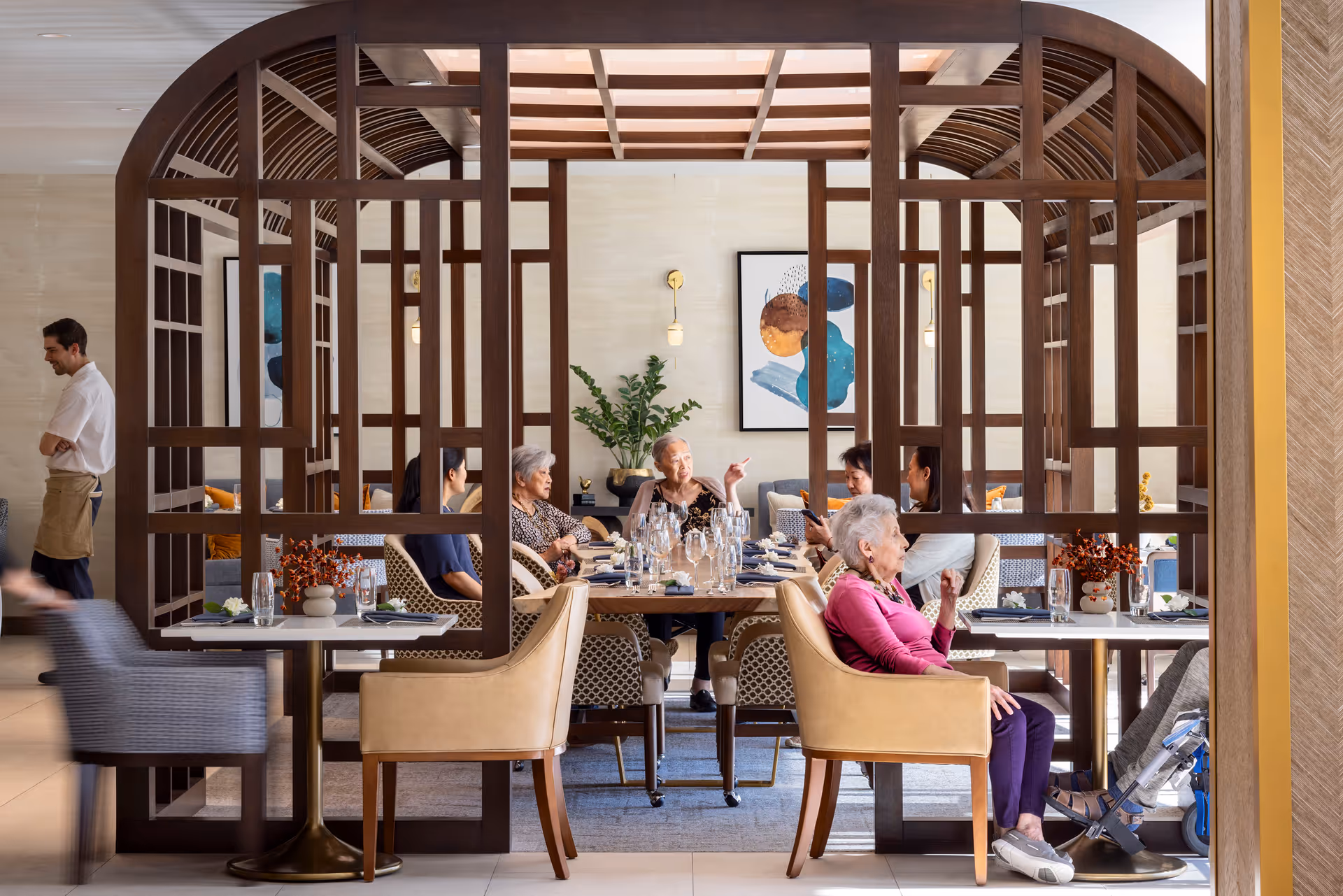 A group of elderly people sitting and conversing around a dining table in a senior living facility dining room. The room features a decorative wooden partition with an arched design, modern chairs, and tables set with glasses and tableware. A staff member stands nearby, and there is artwork and plants on the walls in the background.