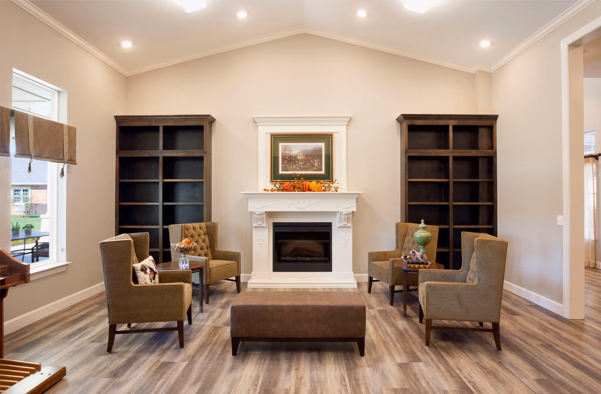 A cozy living room area with four brown upholstered armchairs arranged around a brown ottoman. There is a white decorative fireplace in the center of the back wall with a framed painting above it and autumn-themed decorations on the mantel. Two tall, dark wooden bookshelves flank the fireplace. The room has light-colored walls, wood flooring, and recessed ceiling lights. A window on the left side shows an outdoor view.