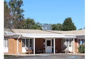 Single-story brick building with a covered entrance and benches outside, surrounded by trees and a clear blue sky.