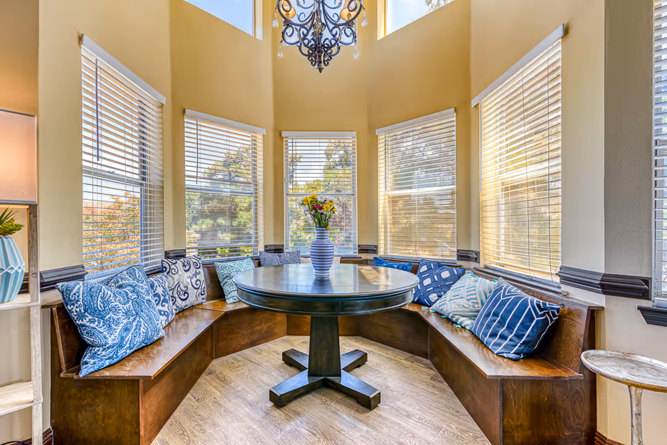 A cozy dining nook with a round wooden table surrounded by a built-in wooden bench adorned with various blue and patterned throw pillows. The nook is enclosed by multiple windows with white blinds, allowing natural light to fill the space. A decorative chandelier hangs from the ceiling, and a vase with flowers sits on the table.