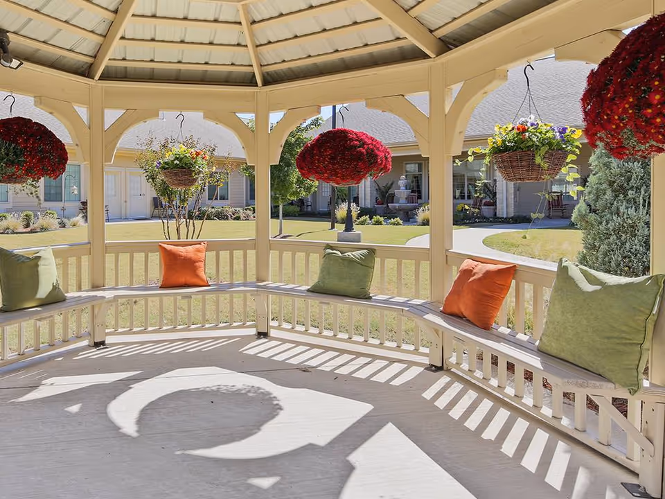 View from inside a gazebo with a curved wooden bench adorned with green and orange cushions. Hanging flower baskets with red and multicolored flowers are suspended from the gazebo roof. The gazebo overlooks a well-maintained lawn and a building with multiple doors and windows in the background.