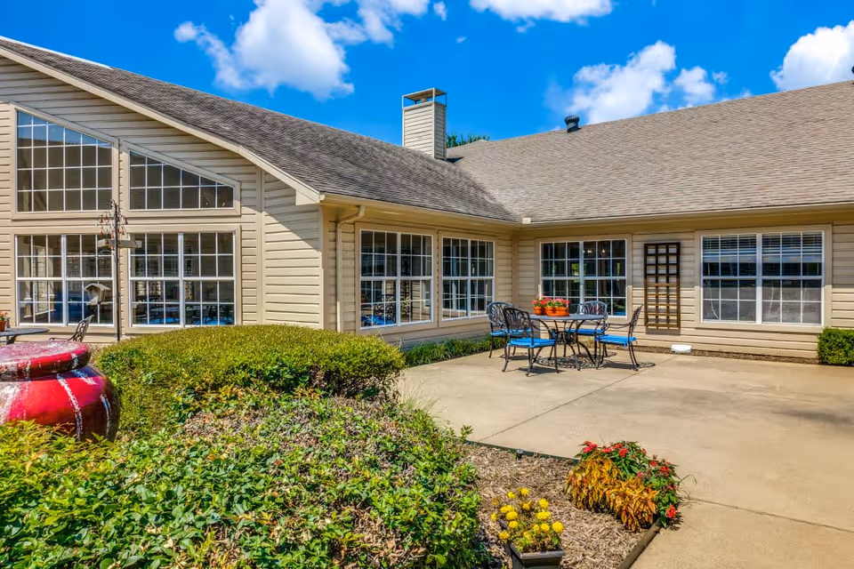 Outdoor patio area of a senior living facility with a concrete floor, metal table and chairs with blue cushions, potted flowers on the table, surrounding bushes and plants, and a beige building with large windows under a blue sky with some clouds.