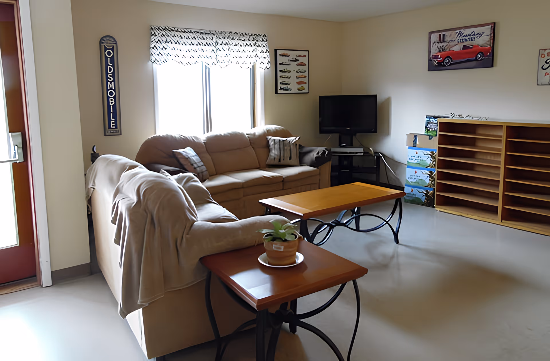 A cozy living room with two beige sofas, a wooden coffee table, and a small side table with a potted plant. A flat-screen TV is placed on a black stand in the corner. The room has a window with a patterned valance, vintage car-themed wall art, and an empty wooden shelving unit against the wall.