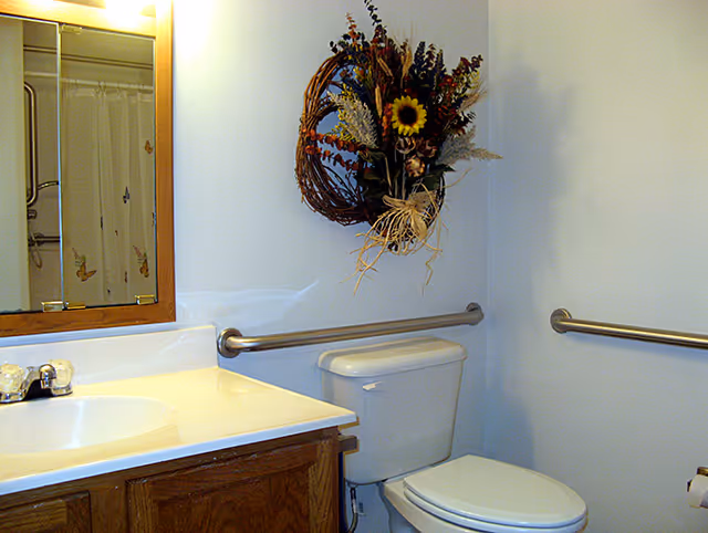 A bathroom with a white toilet and a wooden vanity with a white countertop and sink. Above the toilet, there is a decorative wreath with dried flowers and a sunflower hanging on the wall. There are metal grab bars on the walls near the toilet. A mirror with a wooden frame is mounted above the sink, reflecting part of a shower curtain with butterfly designs.