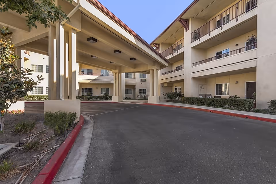 Covered driveway and entrance canopy in front of a three-story beige building with balconies and landscaping.