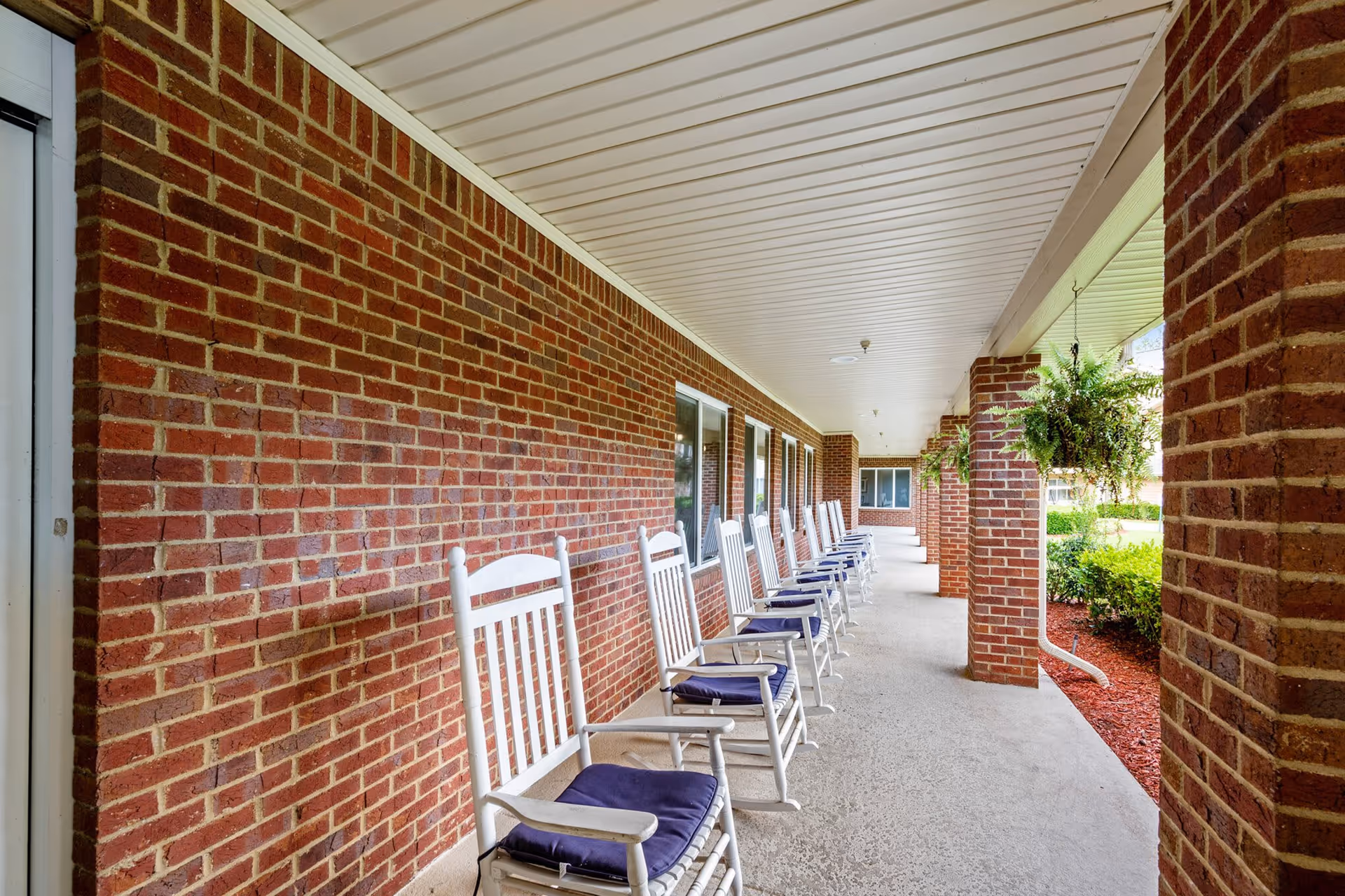 A covered outdoor walkway with a row of white rocking chairs with blue cushions lined up against a red brick wall. Hanging green plants are suspended from the ceiling, and there is a garden area with bushes and mulch visible on the right side.