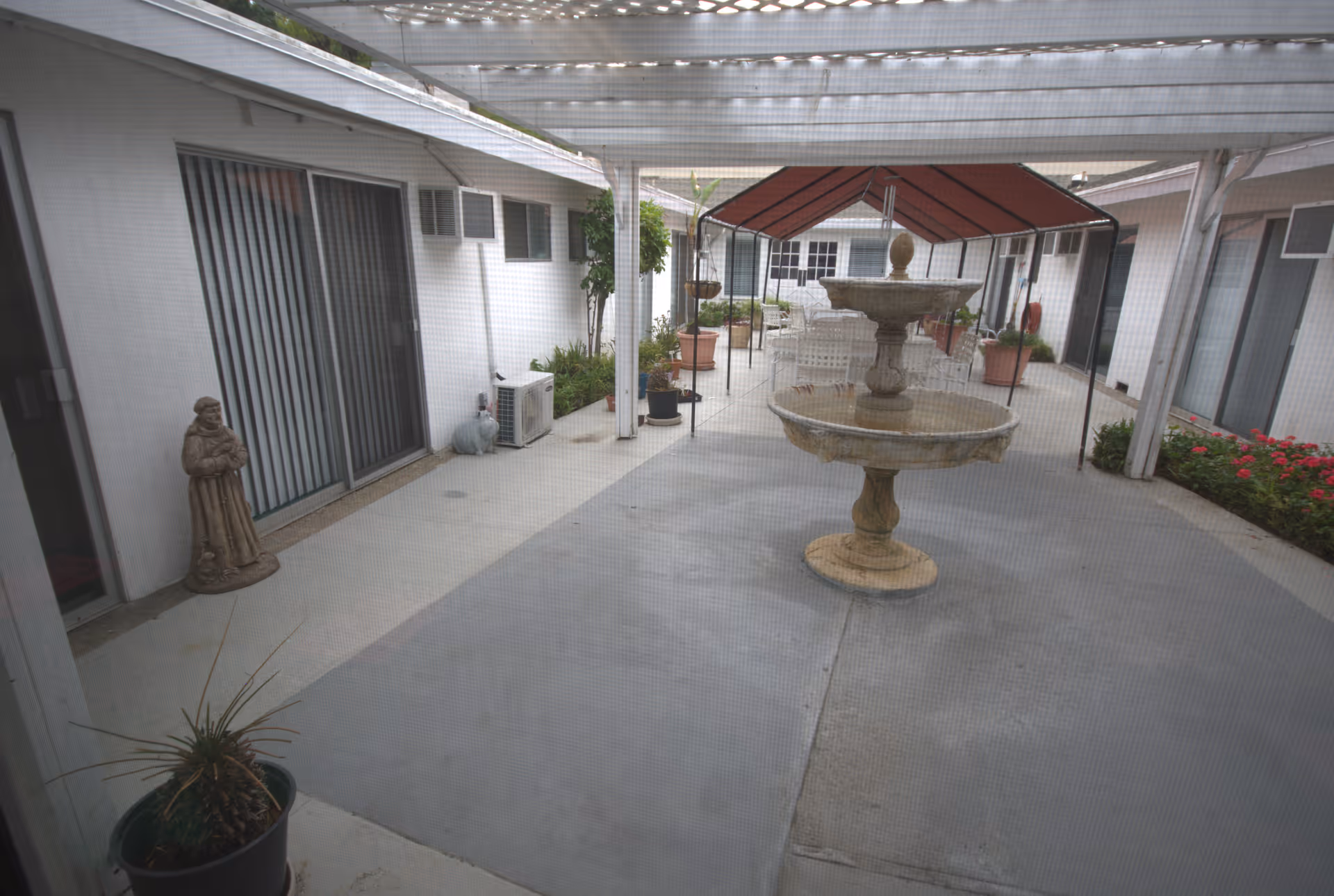 Outdoor courtyard area with a central two-tier stone fountain under a pergola. Surrounding the courtyard are white buildings with sliding glass doors and windows. There are potted plants, a statue, and some greenery along the edges of the courtyard.