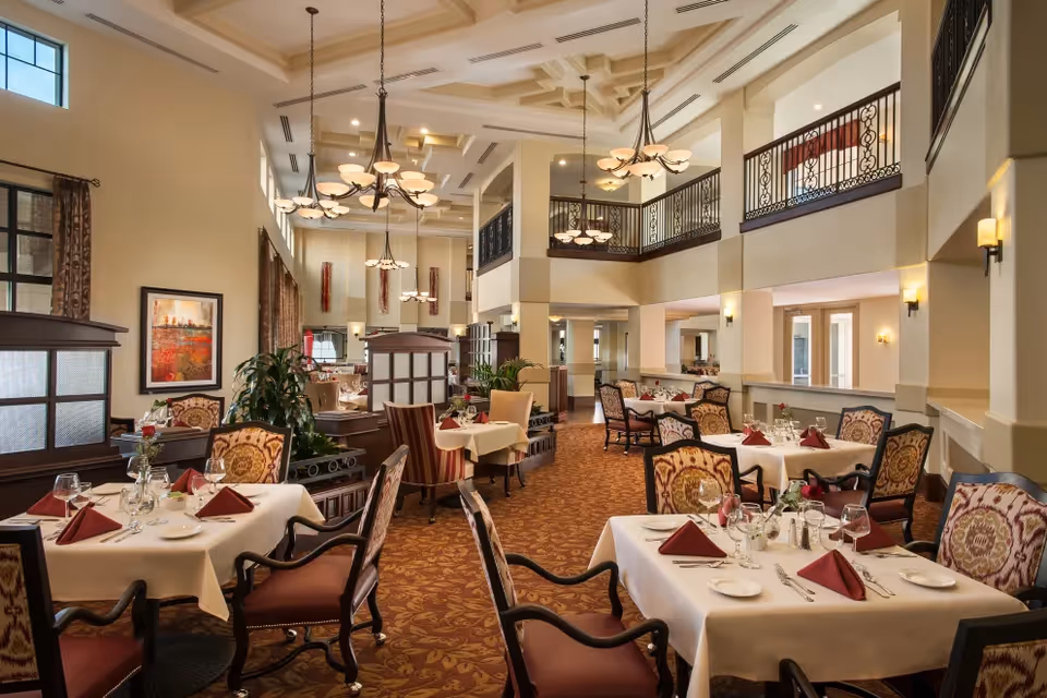 Spacious two-story dining room with neatly set tables, upholstered chairs, chandeliers, and decorative railings.