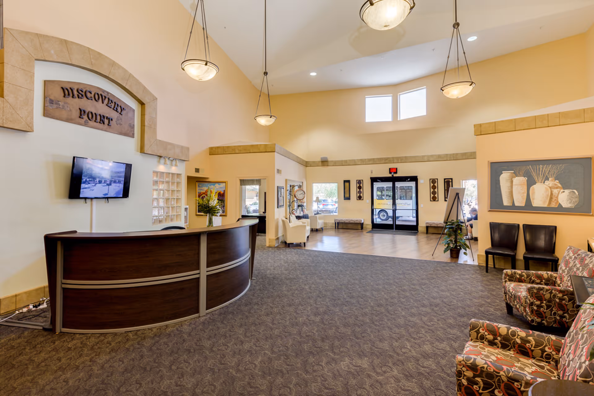 Reception area of Discovery Point Retirement Community with a curved wooden front desk, a wall-mounted TV, comfortable seating with patterned armchairs, decorative vases artwork, and large windows letting in natural light near the entrance doors.