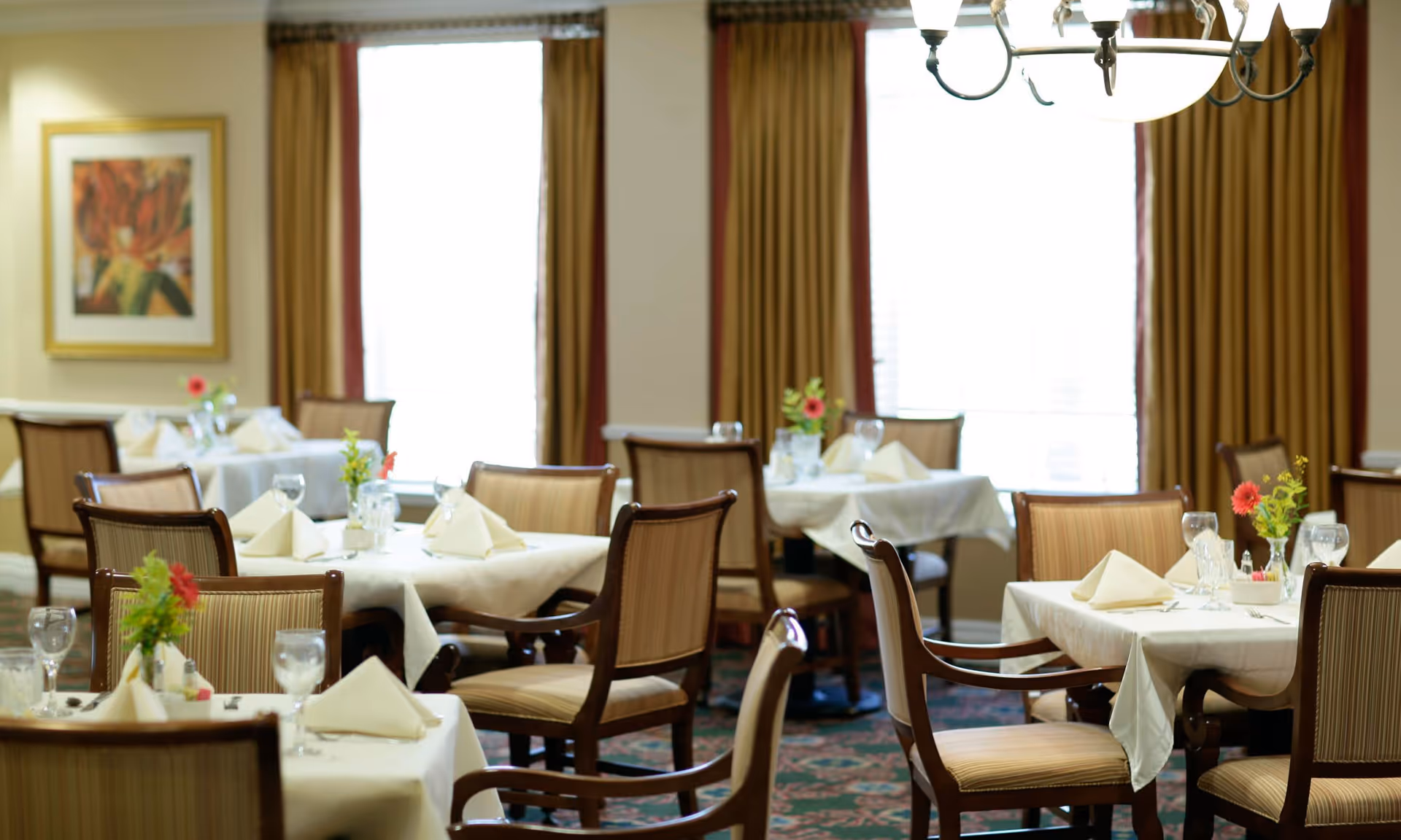 A well-lit dining room with several tables covered in white tablecloths, each set with folded napkins, glassware, and small floral centerpieces. The room has large windows with gold and red curtains, a chandelier hanging from the ceiling, and framed artwork on the wall.